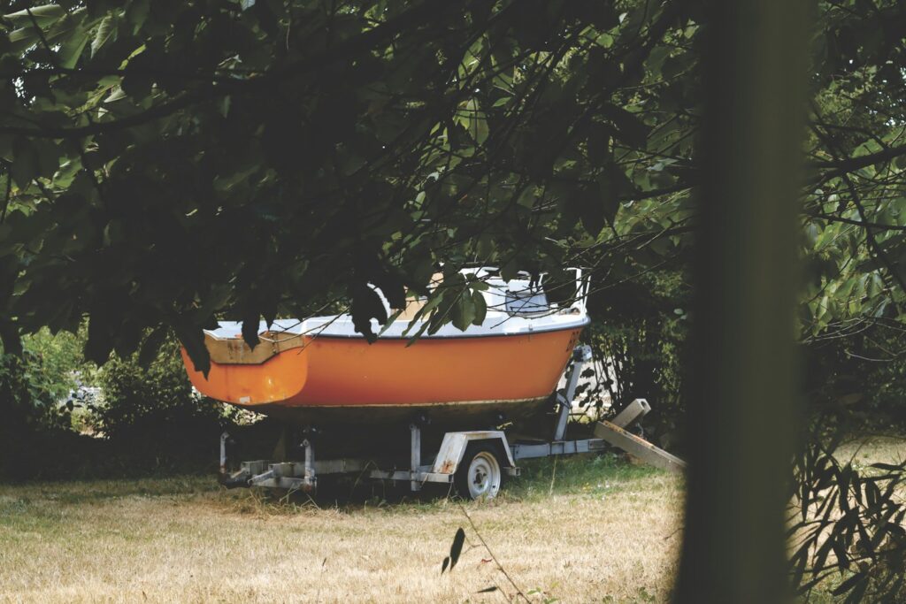 An orange boat on a trailer in a grassy yard under tree branches