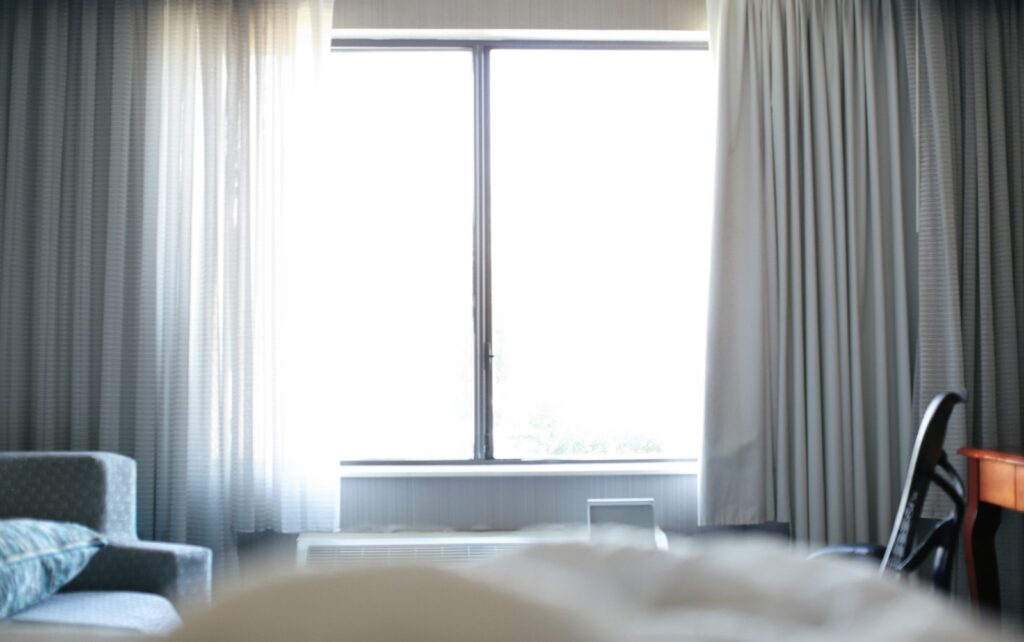 White bed beside a window with white curtains, bringing a light, airy feel to a bedroom